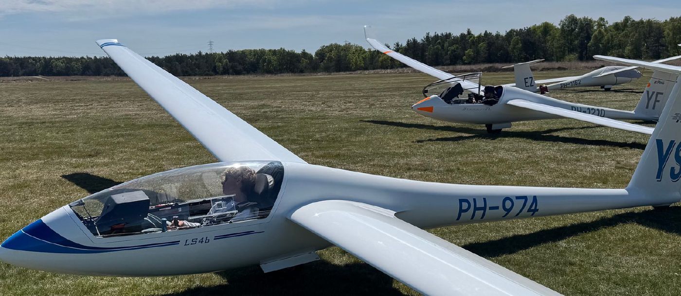 Een lid van ZC Flevo in de cockpit van de LS4-b op het grasveld van Terlet, met andere clubtoestellen en de bosrand op de achtergrond